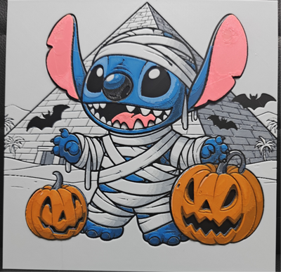 Framed 3D layered art piece featuring a cute illustration of a blue alien character dressed as a mummy wrapped in white bandages, standing between two carved Halloween jack-o'-lantern pumpkins with an ancient Egyptian pyramid and flying bats in the background.