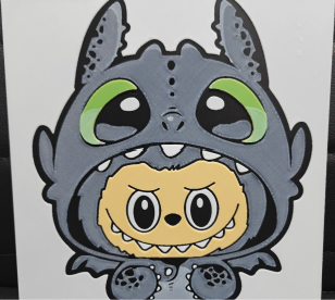 Framed 3D layered art piece featuring a cute illustration of a small cartoon creature with wide eyes and a toothy expression, wearing a dark grey dragon costume hood with large green eyes.