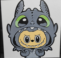Framed 3D layered art piece featuring a cute illustration of a small yellow cartoon creature with wide black eyes and a toothy grin, wearing a textured grey dragon costume hood with large green eyes against a white background.