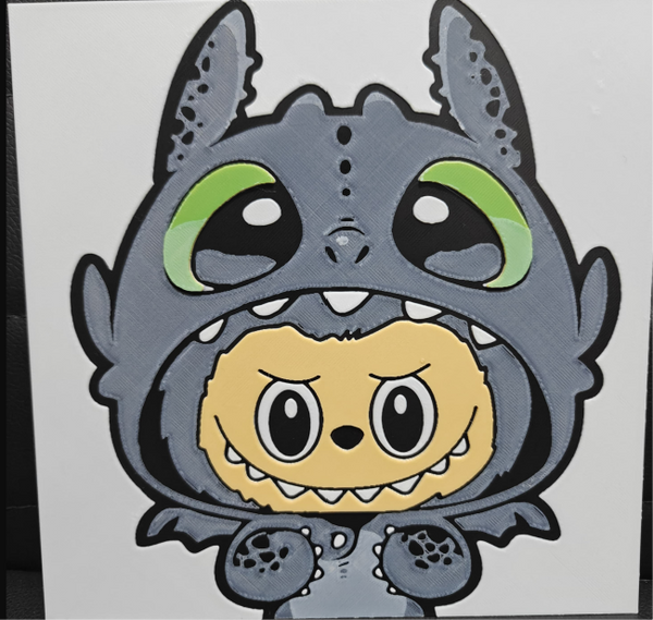 Framed 3D layered art piece featuring a cute illustration of a small yellow cartoon creature with wide black eyes and a toothy grin, wearing a textured grey dragon costume hood with large green eyes against a white background.