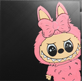 Framed 3D layered art piece featuring a cute illustration of a small cartoon creature with sparkling black eyes and a wide toothy grin, wearing a fuzzy pink costume hood with long ears and a large matching pink bow against a black background.