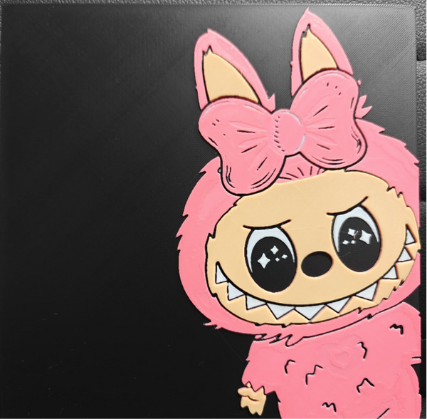 Framed 3D layered art piece featuring a cute illustration of a small cartoon creature with sparkling black eyes and a wide toothy grin, wearing a fuzzy pink costume hood with long ears and a large matching pink bow against a black background.