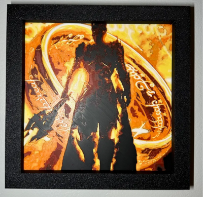 Framed 3D layered art piece featuring a dramatic silhouette of a dark armored fantasy lord holding a large weapon, standing in front of a massive glowing golden ring inscribed with elegant mystical script. The background features an intense, explosive atmosphere of fiery orange lava, flames, and flying embers inside a black square frame.