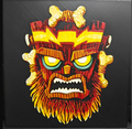 Framed 3D layered art piece featuring a bold illustration of a menacing tribal tiki mask with glowing yellow eyes, fiery orange accents, and sharp geometric details set against a dark background.