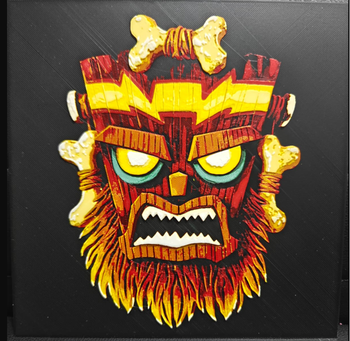 Framed 3D layered art piece featuring a bold illustration of a menacing tribal tiki mask with glowing yellow eyes, fiery orange accents, and sharp geometric details set against a dark background.