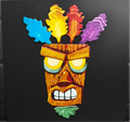 Framed 3D layered art piece featuring a detailed illustration of an iconic gaming wooden witch doctor mask character. The tiki-style mask is shown with large glowing yellow eyes, a determined expression, a green leaf goatee at the bottom, and four colorful upright feathers in blue, yellow, red, and purple across the top against a solid black background.