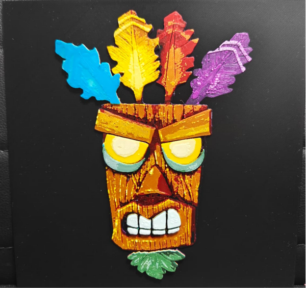 Framed 3D layered art piece featuring a detailed illustration of an iconic gaming wooden witch doctor mask character. The tiki-style mask is shown with large glowing yellow eyes, a determined expression, a green leaf goatee at the bottom, and four colorful upright feathers in blue, yellow, red, and purple across the top against a solid black background.