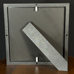 Rear easel view of a WTx3D Large charcoal matte frame with the built-in easel stand open on a wooden surface. Available as Small or Large Framed 3D Layered Art — Large features approximately 8" × 8" artwork in a finished frame measuring approximately 11½" × 11½".