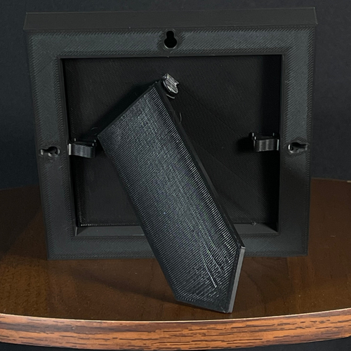 Rear easel view of a WTx3D Small charcoal matte frame with the built-in easel stand open on a wooden surface. Available as Small or Large Framed 3D Layered Art — Small features approximately 4" × 4" artwork in a finished frame measuring approximately 5⅛" × 5⅛".